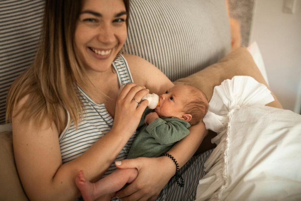 A mother lovingly feeds her newborn with a bottle while smiling at the camera.